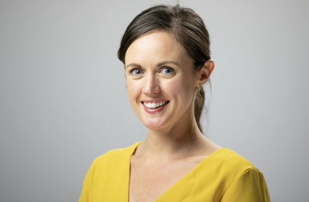 A woman with brown hair wearing a bright yellow shirt smiles at the camera